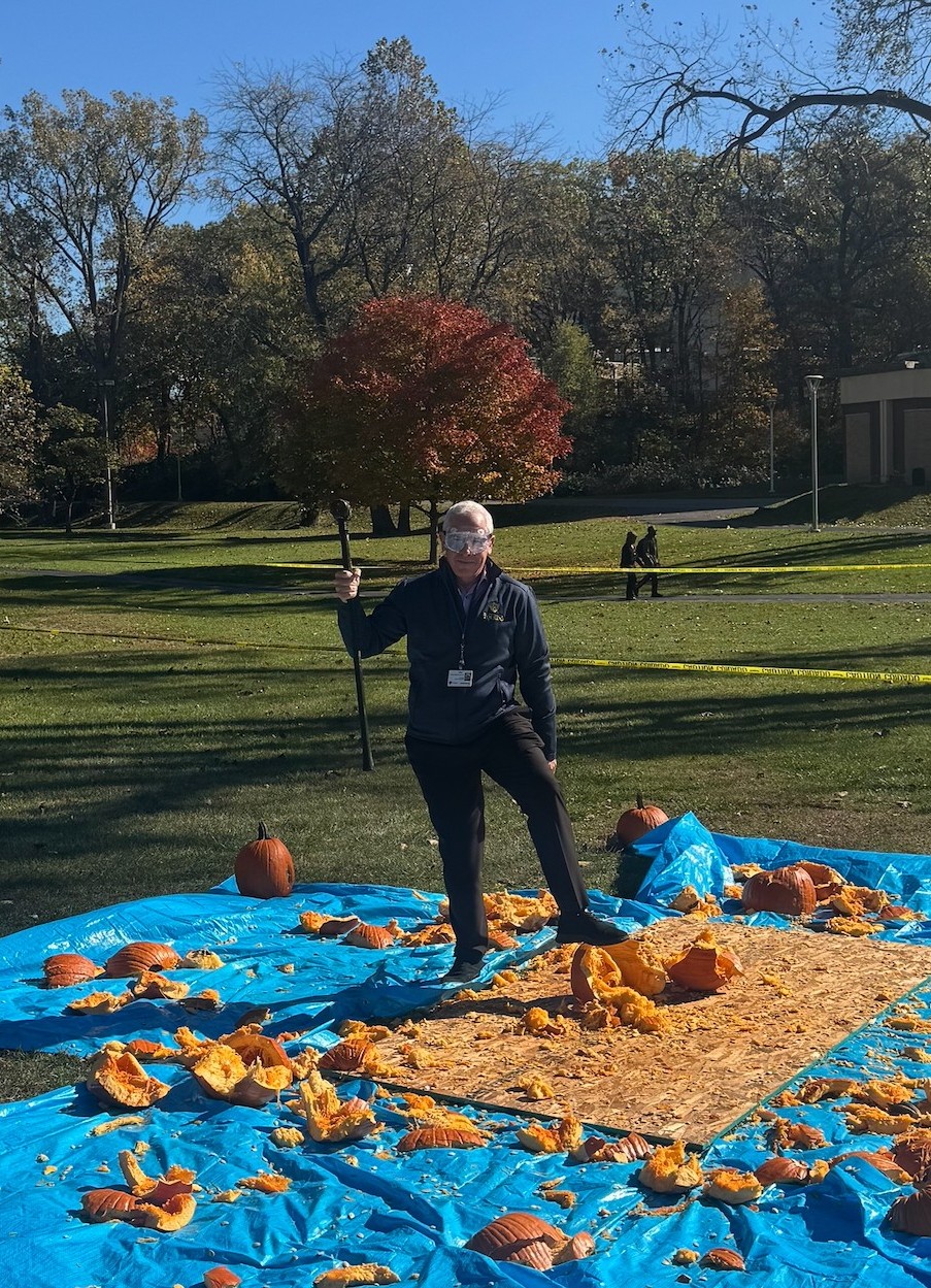 UT President, Dr. Holloway, is seen standing over the remains of several smashed pumpkins. The pumpkin remains are covering four blue tarps and a 8x4 foot particle board. Dr. Holloway is holding up the sledgehammer with his right hand and wearing safety goggles. The large field used for the event is in the background, with a bright blue sky overhead.