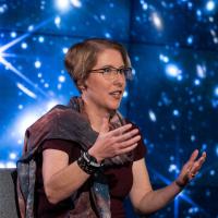 A woman with short hair and glasses gestures while talking in front of an astronomical image